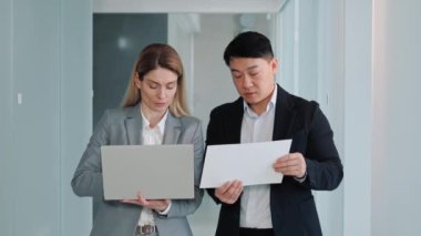 Multicultural business partners working together looking at laptop screen. Two business people working in office hallway reading and discuss asian investment project, analyze the stock market