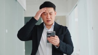 Annoyed angry disappointed male asian bisinessman hold mobile phone reads bad news. Stressed anxious upset holding smartphone thinking about problem solution, loan debt, dismissal, staff cuts notice