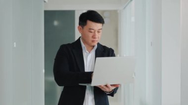 Handsome korean man working on laptop project in office hallway. Online business negotiations. Serious businessman chatting with foreign partners, writing message, look to window, thinking new idea