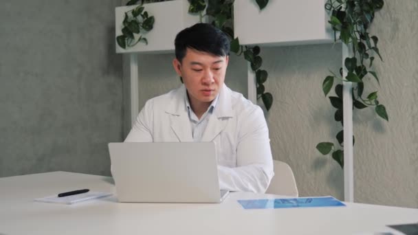 アジアの男性一般開業医は遠く離れた場所でビデオ通話アプリを屋内で使用する患者に医療サポートを提供し クライアントにコンピュータ画面の話を見て距離スピーチを行います 遠隔医療相談の考え方 — ストック動画