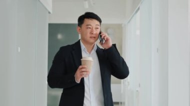 Korean handsome businessman is discussing talking on mobile phone with a pleased smile holdilg coffee standing in the office hall. Successful confident business man having friendly conversation.