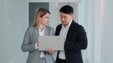 Successful Businesswoman in Stylish Suit Working with Asian Male Adviser. Cheerful Positive Friendly Businesspeople Partners Colleagues Holding Laptop Discuss Ideas. Good Business Relationships