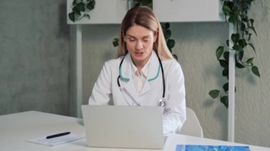 Pretty woman medic looking at screen having online conversation with patient. Caucasian young beautiful female physician in white gown siting at table in cabinet and working at laptop computer