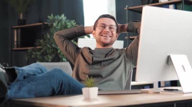 Smiling satisfied young business man freelancer in glasses finished computer work stretching sitting at workplace desk. Happy relaxed businessman resting after job well done putting hands behind head