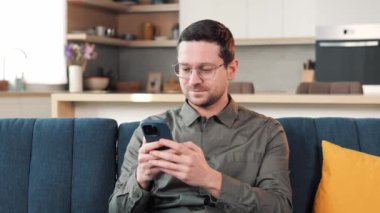 Happy Handsome Caucasian Man in Eyeglasses Using Smartphone in Cozy Living Room at Home. Man Resting on Comfortable Sofa. Browsing Internet, Checking New Videos on Social Networks Typing Message