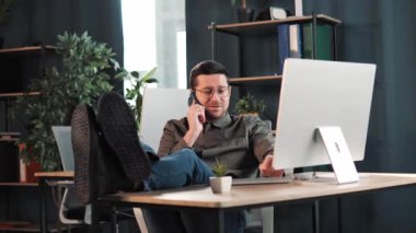 Calm relax free man put legs on table leaning on chair having mobile phone coversation calling enjoy day resting in modern office room looking at laptop screen, no stress, finish work, free time