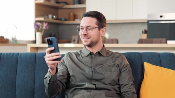 Pozytywnie Przystojny Młody Stylowy Mężczyzna Siedzący Kanapie Salonie Korzystający Smartfona — Wideo stockowe