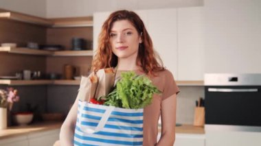 Smiling confident young prettyred hair woman looking at camera showing ok standing alone at home in apartment kitchen. Happy beautiful single millennial lady with a package of eco products, portrait