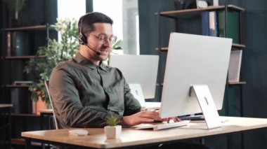 Kulaklık ve gözlüklü genç pozitif adam, internet konferansında internet kamerasından müşteriyle iletişim kurarak iletişim kuruyor. Kulaklık mesafesi ofis sohbetini e-öğreniyor.