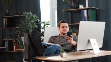Happy rahatla yakışıklı patron yönetici ofis masasında oturmuş cep telefonu kullanarak bilgisayar ekranına bakarak bacaklarını masaya koyuyor. Tüccar hisse senedi alım pazarlığı yapıyor