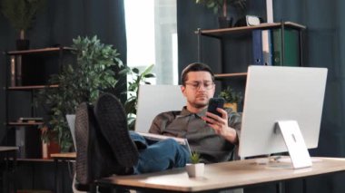 Happy relax handsome male boss leader manager sitting at office desk using mobile phone while typing message looking at pc screen putting legs on the table. Trader negotiates purchase of securities