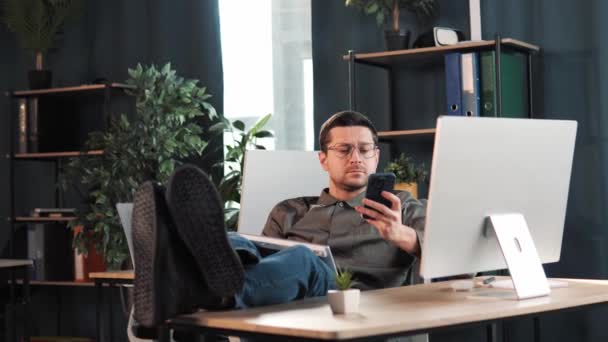 快乐放松英俊的男性老板领导经理坐在办公桌前 一边用手机打字一边看着电脑屏幕 一边把腿放在桌子上 交易者通过谈判购买证券 — 图库视频影像