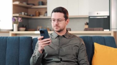 Happy Handsome Caucasian Man in Eyeglasses Using Smartphone in Living Room at Home. Man Resting on Comfortable Sofa Browsing Internet Reading Mail, Checking Videos on Social Networks and Having Fun
