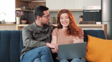 Cheerful Couple Use Laptop Computer, Sitting on Cozy Sofa in Living Floor room of their Apartment. Boyfriend and Girlfriend Talk, Shop on Internet, Choose Product to Order Online, Planning a Vacation