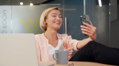 Ofiste cep telefonuyla konuşan gülümseyen bir kadın iş kahkahalarını arkadaşlarıyla konuşurken tartışıyor. Uzaktan iletişim yetenekleri. Video çağrısı, uzaktan çalışma, online iletişim kavramı