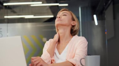 Yorgun ve aşırı çalışan sarışın iş kadını modern bir ofiste iş yerinde oturuyor, uzun çalışma saatlerinden dolayı boynu ağrıyor. Dizüstü bilgisayarda bir tasarım projesi üzerinde çalışan bir kadının dinlenmeye ihtiyacı var