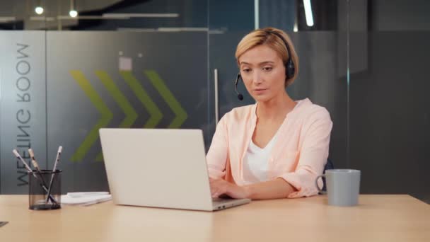 コールセンターで働くヘッドセットを身に着けている明るいオフィスの設定の若い女性は サポートを提供する顧客と話します 女の子はノートパソコンと机に座って 彼女の仕事に焦点を当て表示されます — ストック動画