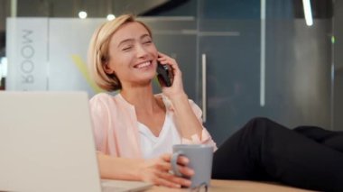 Ofisteki zeki sarışın kadın telefonla konuşuyor kahve içiyor, muhtemelen iş arkadaşı ya da müşterisiyle. Kız gülümsüyor ve sohbet sırasında mutlu görünüyor, başarılı bir proje ya da anlaşma tartışması yapıyor.
