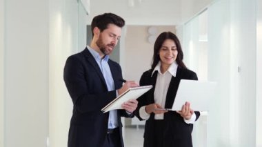 Dizüstü bilgisayarları ve not defterleri olan iki çalışanı ofis koridorundaki yeni proje üzerinde işbirliği yapıyor, fikirleri paylaşıyor ve bunların yaratıcılığından faydalanıyor. Ekranda bilgi gösteren kadın ofis çalışanı