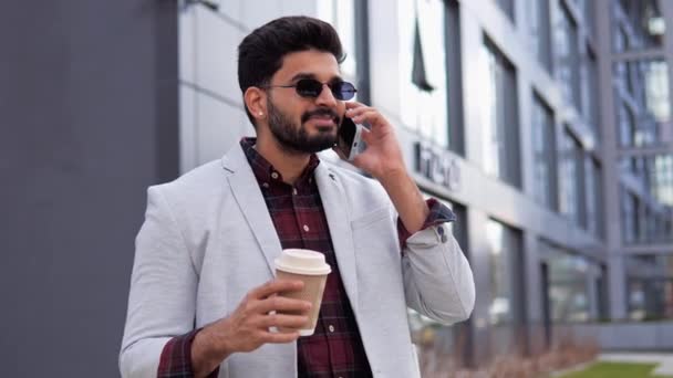 精力充沛的印度混血儿商人戴着太阳镜 拿着一杯咖啡 穿着时髦而凉爽的衣服 一边用手机聊天 商人在手机上讲话 — 图库视频影像