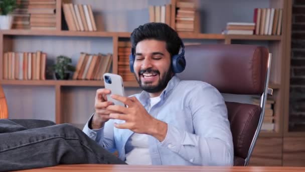 職場に座っている面白い若いインドの男性会社の代表取締役は ワイヤレスヘッドフォンで好きな音楽を聞いています 幸せなリラックスしたビジネスマン休憩時間を楽しんで オフィスで歌を歌う — ストック動画