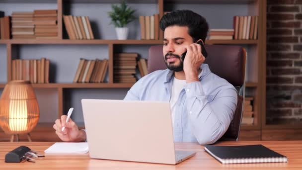 彼のオフィスでラップトップを使用している間 インドのビジネスマンは喜んで彼の携帯電話で会話します 男は仕事とコミュニケーションをシームレスに管理し マルチタスクに表示されます 男性用筆記体 — ストック動画
