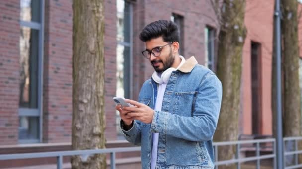 オフィスビルの近くの屋外を歩いてスタイリッシュなデニムジャケットを身に着けているインドの成功したビジネスマンの肖像画は 考えて検索メッセージを入力します スマートフォンを屋外に持ちながら笑う男 — ストック動画