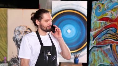 Yakışıklı erkek sanatçı, stüdyosunda elinde cep telefonuyla soyut tablolarının önünde duruyor. Beyaz tişörtlü bir adam işiyle ilgilenirken cep telefonuyla konuşuyor.
