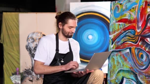 一位有魅力的男性艺术家穿着一件白色的T恤 正在工作室里画一幅画 人们一边看抽象画 一边用手提电脑和键盘打字 描述了他们的销售情况 — 图库视频影像