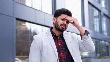 Hintli bir adam bir binanın önünde dikilirken stres ve baş ağrısı yaşıyor. Finansal bir sorun ya da iş başarısızlığı hakkında endişeli görünüyor. Zihinsel sağlık sorunları, iş stresi, mali zorluklar