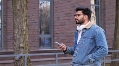 Kulaklıklı yakışıklı bir Latin adam sokakta yürüyor, cep telefonuyla sohbet ediyor, kaydırırken mesaj yazıyor. Sosyal medyada sörf yapıyor, boş zamanlarının ve tatil konseptinin tadını çıkarıyor.