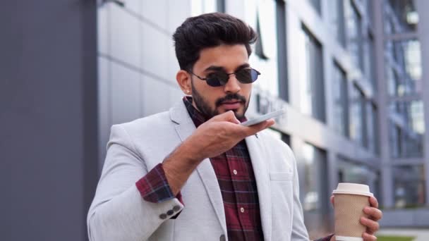 身穿黑色太阳镜的时髦 酷酷的印度人一边在街上端着一杯咖啡 一边拿着手机记录着一条语音留言 与手机管理员交谈 使用智能手机语音识别的人 — 图库视频影像