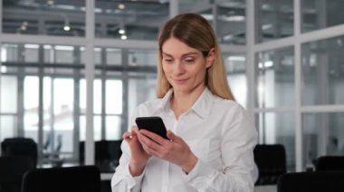 Güzel görünümlü bir iş kadını, sosyal medya ile iletişim kurarken yüzünde mutlu bir gülümsemeyle, cep telefonuna mesaj yazarken ofis elbisesi içinde görülüyor. Kadın kapalı alanda cep telefonu üzerinde çalışıyor.