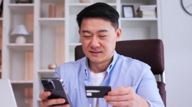 Mavi gömlekli gülümseyen Çinli işadamı, elinde kredi kartı ve cep telefonu olan bir masada oturuyor ve e-ticaret için CVV kodu giriyor. Odaklanmış Asyalı adam alışveriş yapıyor.