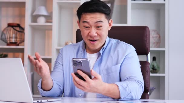 青いシャツを着たアジアのビジネスマンがオフィスの机に座っていて 良いニュースを祝うスマートフォンを持っています 男性はモバイルオンラインベットを獲得し 金融賞を受賞しました — ストック動画