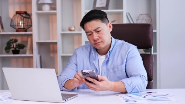 人们看到疲惫 精疲力竭的亚洲男性办公室职员坐在办公室的办公桌前 拿着手机 翻阅社交媒体的新闻资料 对工作不感兴趣 疲惫不堪 — 图库视频影像