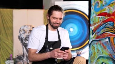 Uzun saçlı, beyaz tişörtlü ve siyah boyalı önlüklü mutlu bir ressam, elinde bir cep telefonu tutuyor ve arka planda resimlerle sosyal medyada sohbet ediyor.