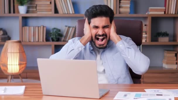 青いシャツを着たインド人ビジネスマンが彼のオフィスに座って仕事をしようとしています 人間は外部の騒音や断熱材に気を取られ 耳を手で覆い 屋内で叫ぶ — ストック動画
