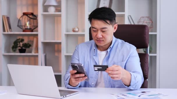 有魅力的亚洲男人面带笑容 穿着蓝色衬衫 坐在办公室的办公桌前 手里拿着手机和信用卡 小心翼翼地输入Cvv代码 完成了电子商务的采购 — 图库视频影像
