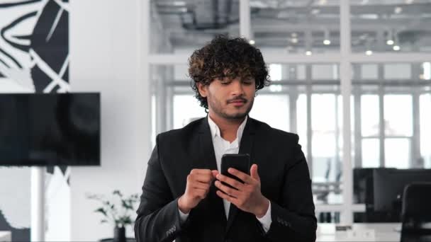 現代のオフィスで携帯電話のアプリを使用して自信を持って男 インドのビジネス専門家は財務ニュースを閲覧し 笑顔は良いニュースを読みます 成功した男性は最新のニュースやトレンドで最新の状態に滞在します — ストック動画