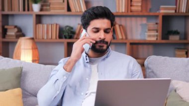 Kanepede oturmuş akıllı telefondan konuşan pozitif gülümseyen Hintli adam. Sohbet etmek için cep telefonu kullanan mutlu erkek işadamı. Keyifli adam iletişim için telefon kullanıyor.