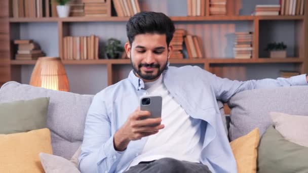 目元居心地の良いリビングルームで快適なソファでリラックスしながら スマートフォンを使用しているインド人男性 リラックスした男はインターネットを閲覧し ソーシャルネットワークでビデオをチェックし 自宅で楽しい時間を過ごしています — ストック動画