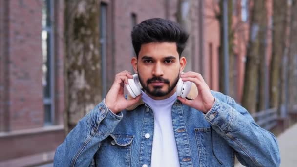 流行のファッショナブルなインド人男性は 屋外で笑みを浮かべてカメラを探して白いヘッドフォンに置くジーンズのジャケットを着て スマートフォンで音楽アプリを使う男今日の若者文化における娯楽 — ストック動画