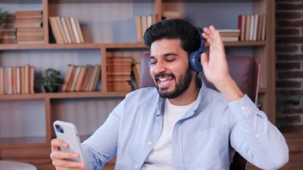 職場に座っている陽気な若いインドの男性会社の代表取締役は ワイヤレスヘッドフォンで好きな音楽を聞いています 幸せなリラックスしたビジネスマンオフィスで休憩時間の歌を楽しんで — ストック動画