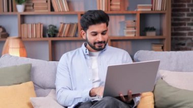 Bin yıllık ciddi Hintli etnik erkek serbest öğrenci ofis koltuğunda dizüstü bilgisayar kullanıyor, apartmanda uzaktan not defteri üzerinde çalışan karışık ırk girişimcilerine odaklanıyor.