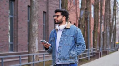 Kulaklıklı yakışıklı Latin bir adam sokakta yürüyor, cep telefonuyla sohbet ediyor, kaydırırken mesaj yazıyor. Sosyal medyada sörf yapıyor, boş zamanlarının ve tatil konseptinin tadını çıkarıyor.