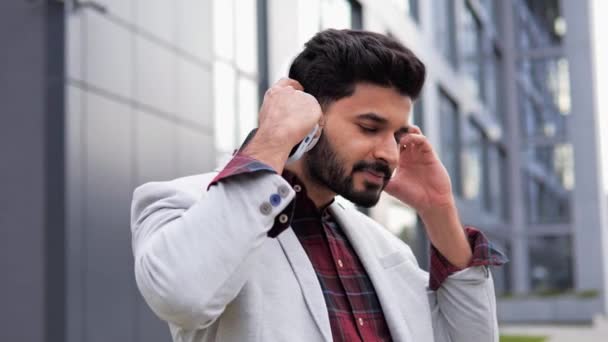 幸せな正のヒンズー系インド人の男のフリーランサーはヘッドフォンを取る 笑顔の男が屋外の都市部の通りに立って カメラを見て 頭からヘッドフォンを取る 男は携帯電話で音楽を聴く — ストック動画