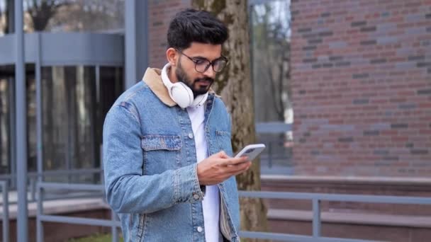 携帯電話のスマートフォンの画面を見て街を歩くメガネの若いビジネスマンの幸せなラテン系のヒスパニック系の男はガジェットを使用してソーシャルメディアでネットの笑顔の答えメッセージでオンラインチャット — ストック動画