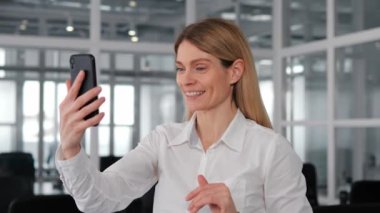 Neşeli iş kadını iş yerinde akıllı telefondan bir video görüşmesi yapıyor. Bayan ofis çalışanı iş arkadaşıyla iletişim kurmak için telefonunu kullanıyor. Kameraya işaret eden, karizma ve coşku saçan bir kadın.