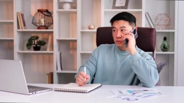 Akıllı Asyalı girişimci dizüstü bilgisayarla çalışırken akıllı telefondan konuşuyor. Ofis masasında oturan kendine güvenen ciddi bir iş adamı. Ceo yöneticisi cep telefonu aramasında not alıyor